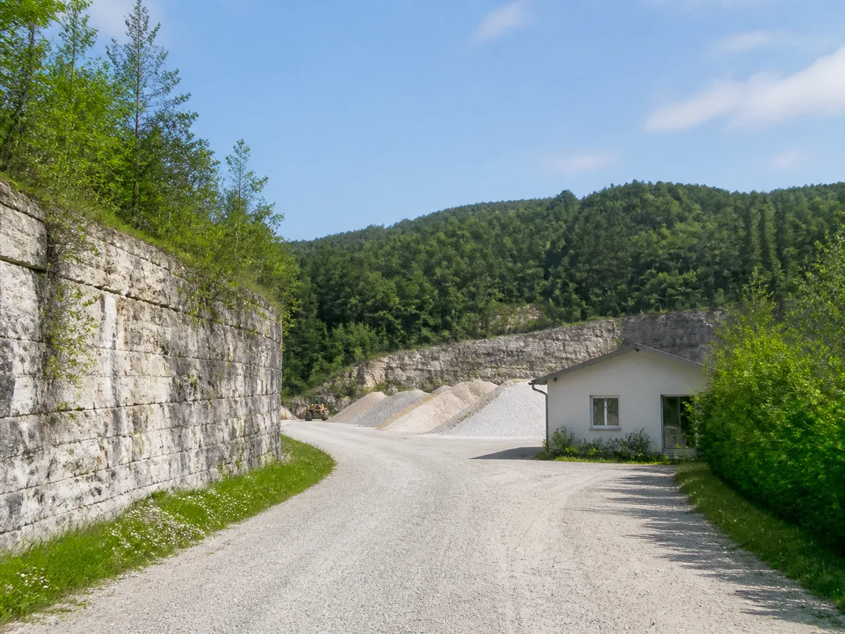 Zufahrt zu einem österreichischen Natursteinstandort mit Kalksteinwand, Grünrand und kleinem Betriebsgebäude